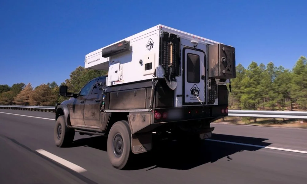 OEV Back Country truck camper mounted on an Aluma Tray flatbed with side storage boxes and a rear box, traveling on the highway with the pop-up roof closed