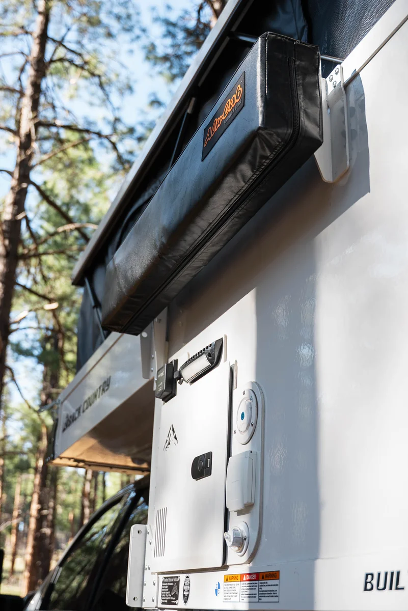 Back Country 6.85 driver-side detail showing awning bracket, Truma Aquago hot water heater access panel, and Back Country badge