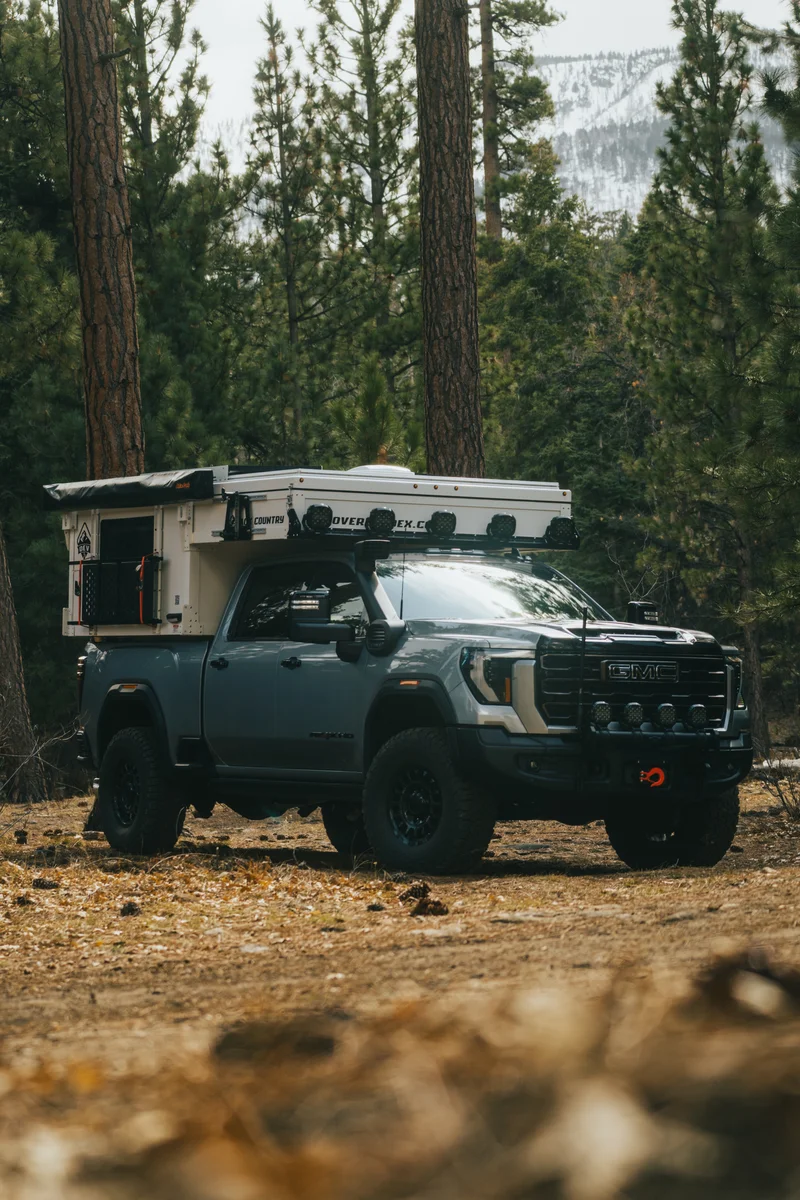 Back Country 6.85 slide-in truck camper on a GMC truck with the roof closed, pine forest and snow-capped mountains behind