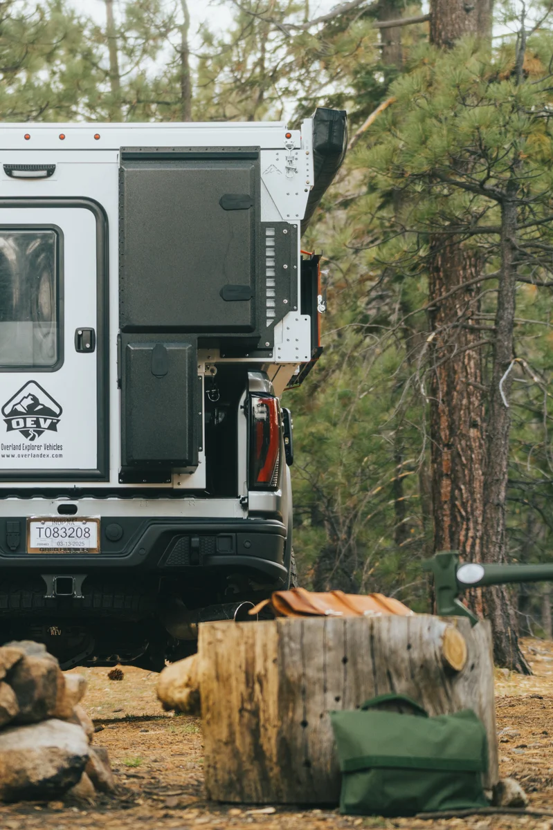 Back Country 6.85 rear three-quarter close-up showing entry door, OEV badge, and camp gear at basecamp