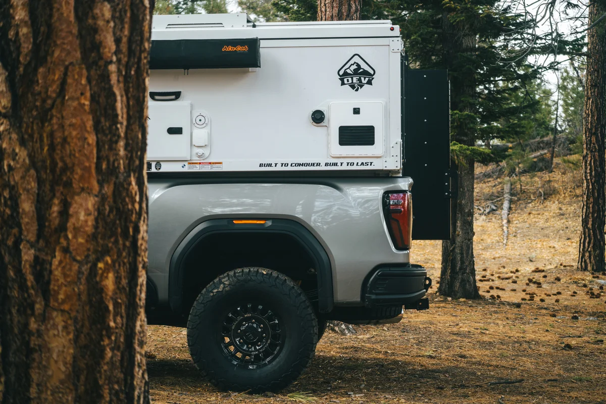 Back Country 6.85 passenger side framed through pine tree showing Alu-Cab awning and 'Built to Conquer' decal