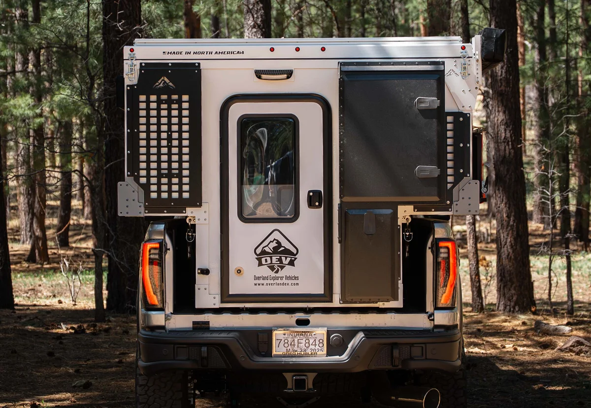 Rear view of a Back Country 6.85 slide-in camper showing the entry door with OEV badge, the MOLLE panel, and the two-door rear storage box