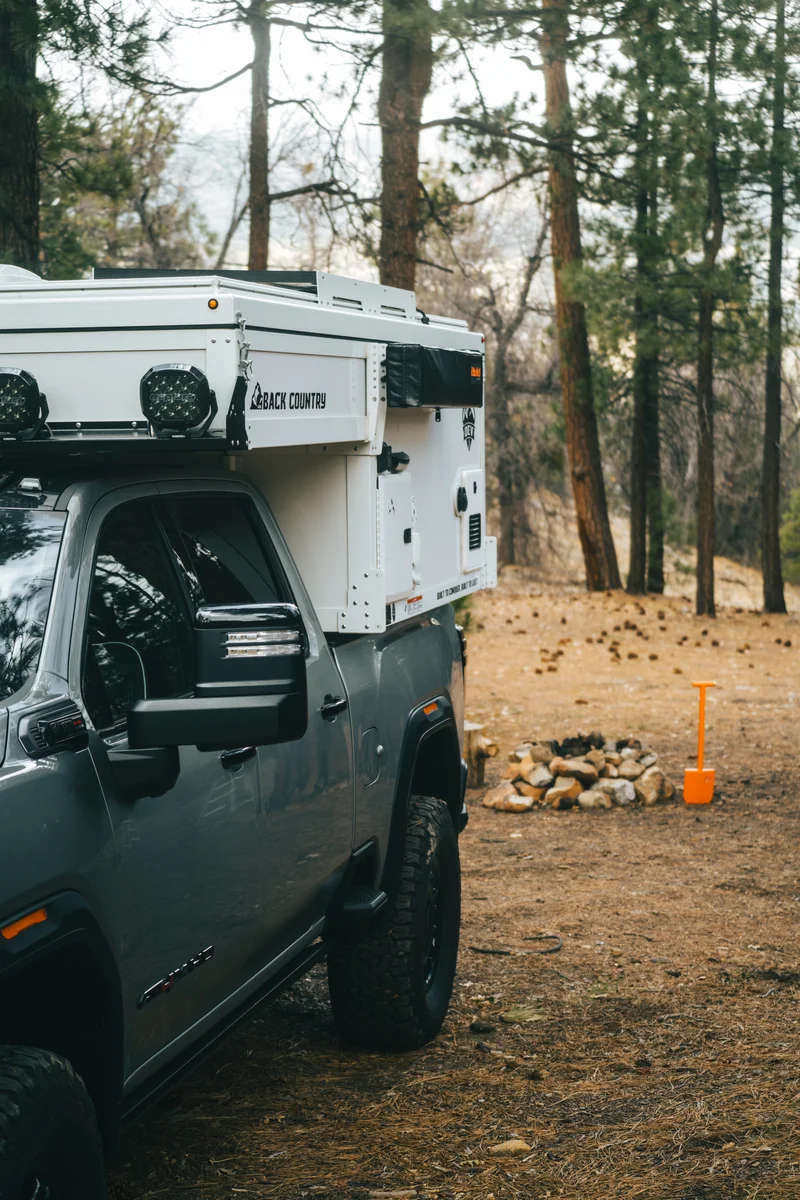 Back Country 6.85 driver side at camp with Back Country badge visible and fireside setup with orange shovel