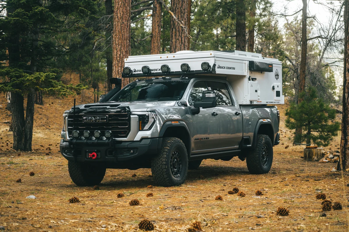 Back Country 6.85 slide-in pop-up camper on a GMC Sierra HD, front three-quarter view with the roof closed and the light bar mounted, in a pine forest