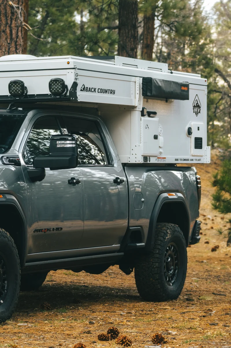 Back Country 6.85 driver side forward quarter portrait showing GMC AT4X HD badge, roof rack, and Back Country camper