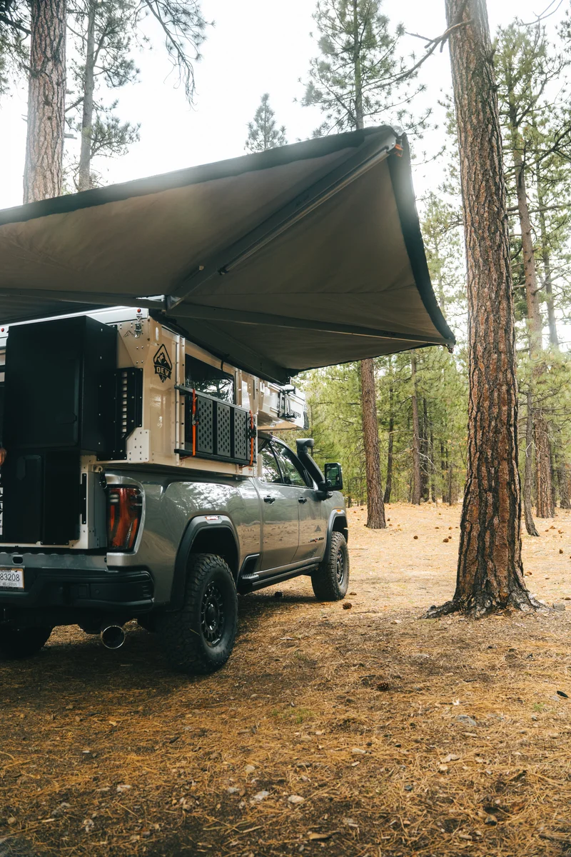 Alu-Cab 270 awning fully deployed from the driver side of a Back Country 6.85 slide-in camper, pop-up roof closed, in a pine forest