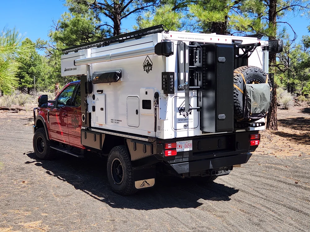 Hudson Bay 8' flatbed pop-up truck camper with the roof closed, rear three-quarter view showing the rear ladder, spare tire carrier, and black flatbed on a red pickup among pine trees
