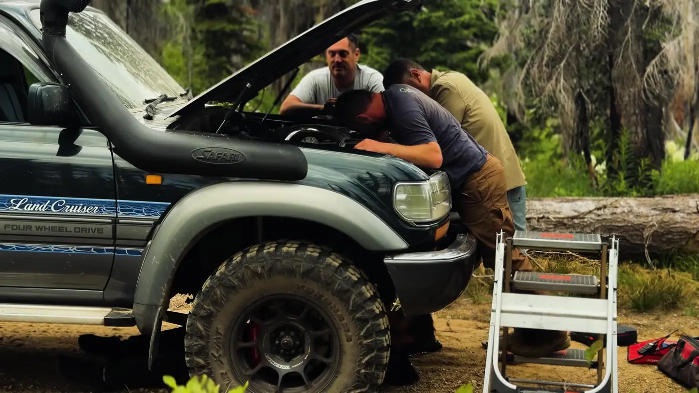Three people leaning under the open hood of a Toyota Land Cruiser on a forest road, with a step ladder set up beside the front tire