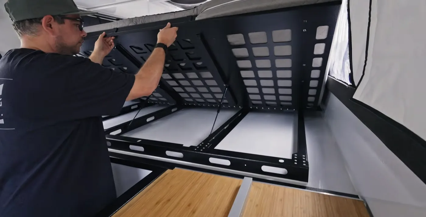 Daniel of Tiny Rig Co. lifting the over-cab bed platform in a Hudson Bay 6.75' to show the under bed storage compartments, slotted aluminum panels, and gas struts