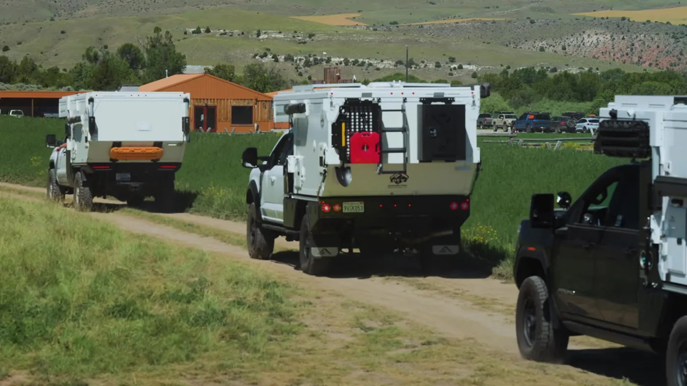 Trucks with white truck campers following a dirt two-track past ranch buildings and green fields at the XOVERLAND Ranch during The Big Thing 2026