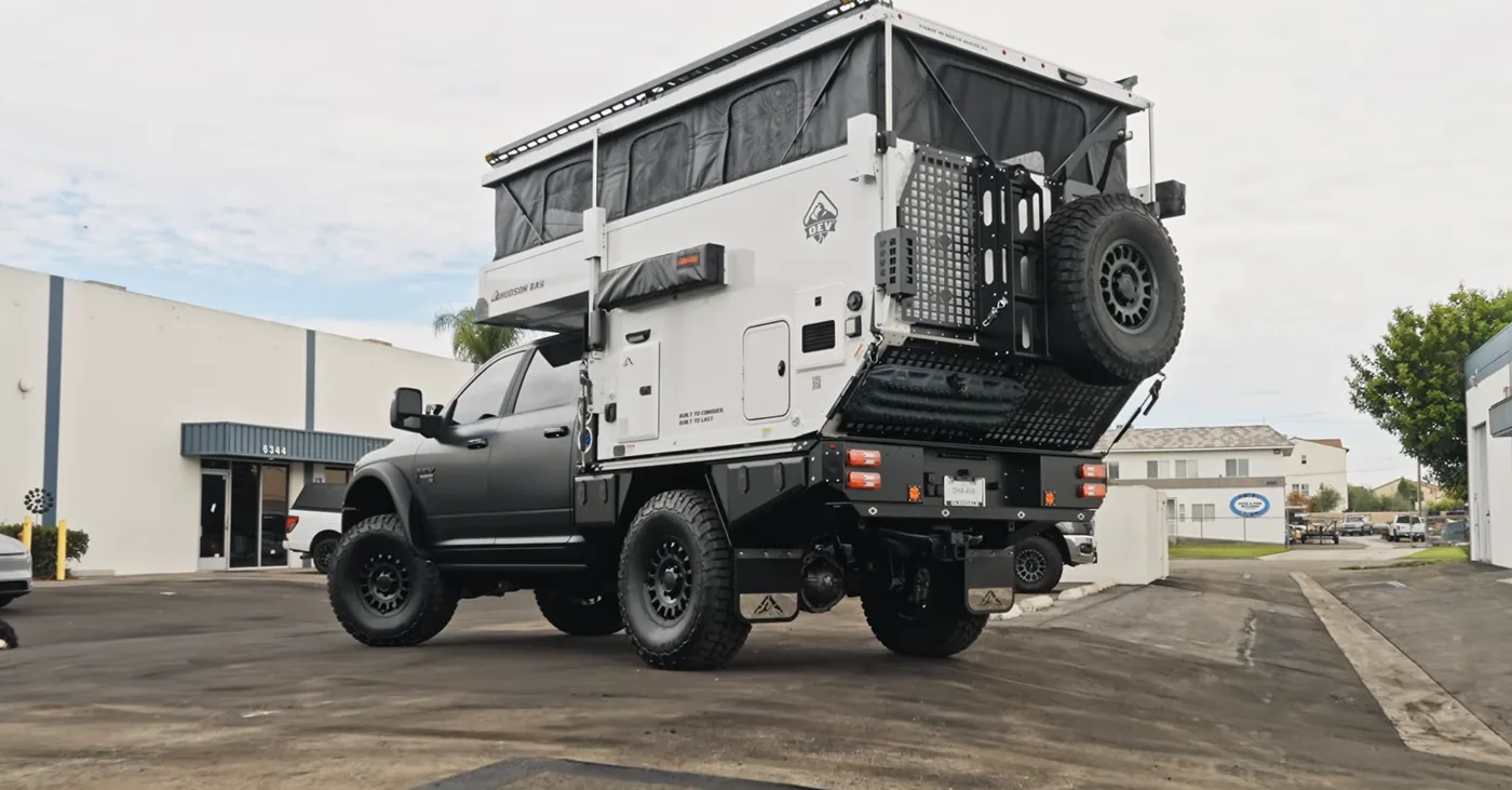 Dark gray Ram 3500 AEV Prospector XL carrying an OEV Hudson Bay 6.75' flatbed pop-up camper on an Aluma Tray, rear three-quarter view with the roof raised, spare tire carrier, and MOLLE panels