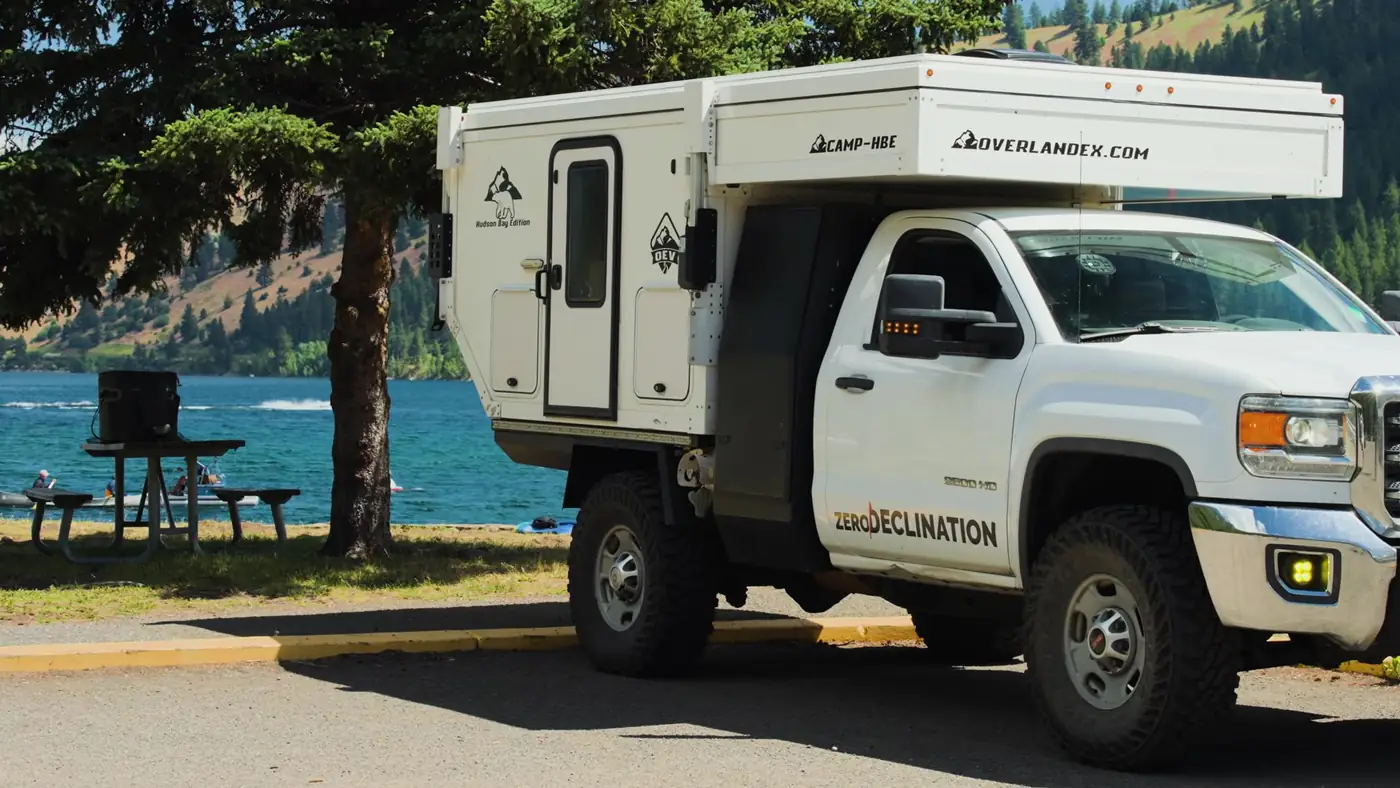 White GMC truck with Zero Declination door lettering carrying an OEV Hudson Bay pop-up camper, parked under pines beside the blue water of Wallowa Lake in Oregon